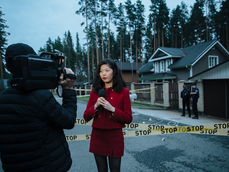 Newscaster reporting on camera beside home surrounded by police officers and caution tape
