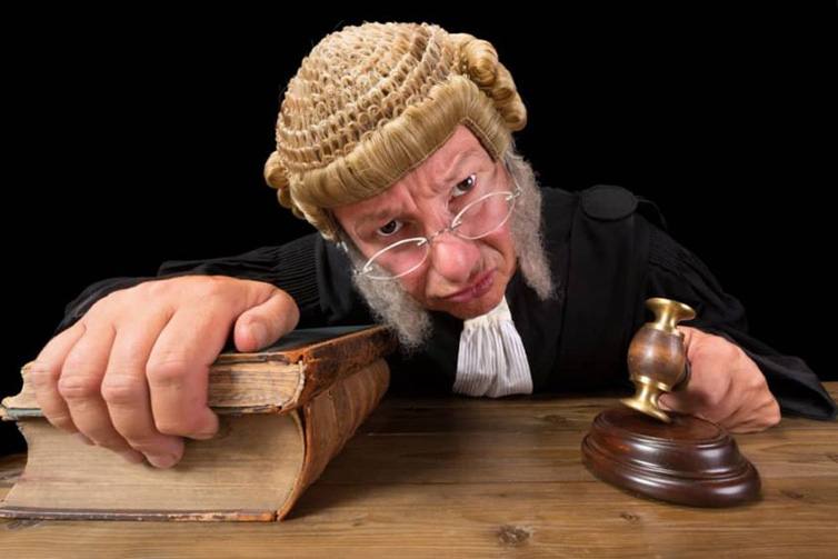 Judge in old-fashioned wig gripping bible and gavel leans forward with menacing expression