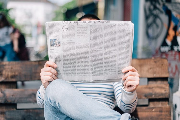 Person reading newspaper in long-sleeved striped shirt and jeans on wooden bench outdoors